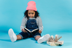 Cute Black girl wearing striped shirt and overalls sits on floor and reads storybooks (Photo: Amina Filkins/Pexels)