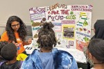 Grade school students stand around a table with a science project on it as a grad student talks about it.