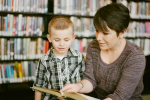 Woman helps young child read a book in the library