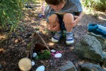 Child kneeling near a small structure on the ground made up of sticks, moss, rocks, and acorns.